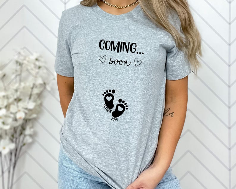 Op de afbeelding: Een lichtgrijs t-shirt met korte mouwen en de zwarte tekst "COMING..." boven "soon", geflankeerd door twee kleine hartjes. Daaronder staat een afbeelding van twee babyvoetjes met hartvormige hielen en de zwarte tekst "OCTOBER 2024".