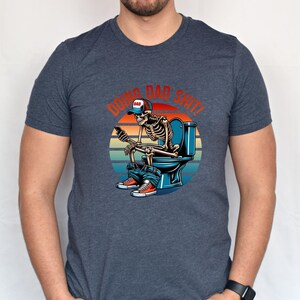 May include: Dark blue t-shirt featuring a skeleton on a toilet, wearing a baseball cap that says "DAD" and holding a phone. The graphic includes the text "DOING DAD SHIT!" in red and orange.