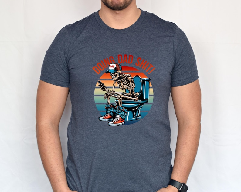 May include: Dark blue t-shirt featuring a skeleton on a toilet, wearing a baseball cap that says "DAD" and holding a phone. The graphic includes the text "DOING DAD SHIT!" in red and orange.