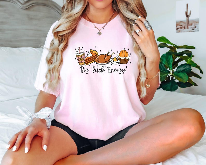 Puede incluir: Camiseta rosa claro con la frase "Big Back Energy" e ilustraciones de un caf&eacute; con leche de calabaza, tarta, pavo y cupcake. La camiseta est&aacute; hecha de un material suave.