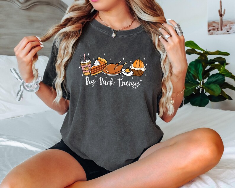 Puede incluir: Camiseta gris oscuro con un gr&aacute;fico de alimentos de Acci&oacute;n de Gracias, que incluye un caf&eacute; con leche de calabaza especiada, pastel, pavo y una magdalena. El texto "Big Back Energy" est&aacute; escrito debajo en blanco.