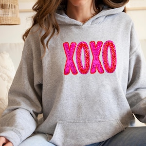 以下が含まれることがあります： 前面にピンクと赤のグリッターのXOXOグラフィックが施されたグレーのパーカー