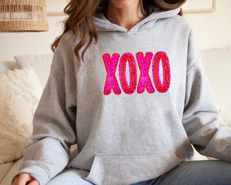以下が含まれることがあります： 前面にピンクと赤のグリッターのXOXOグラフィックが施されたグレーのパーカー
