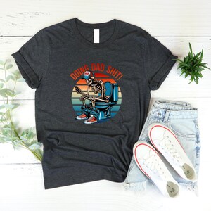 May include: Dark gray t-shirt featuring a skeleton on a toilet with the text "DOING BAD SHIT!" above the skeleton. The skeleton is wearing a baseball cap, jeans, and sneakers. The design includes a sunset background.