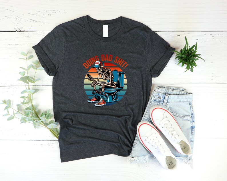 May include: Dark gray t-shirt featuring a skeleton on a toilet with the text "DOING BAD SHIT!" above the skeleton. The skeleton is wearing a baseball cap, jeans, and sneakers. The design includes a sunset background.