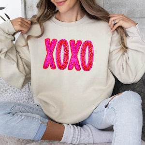 以下が含まれることがあります： クリーム色のスウェットシャツに、ピンクと赤のグリッターで「XOXO」のグラフィックが施されています。