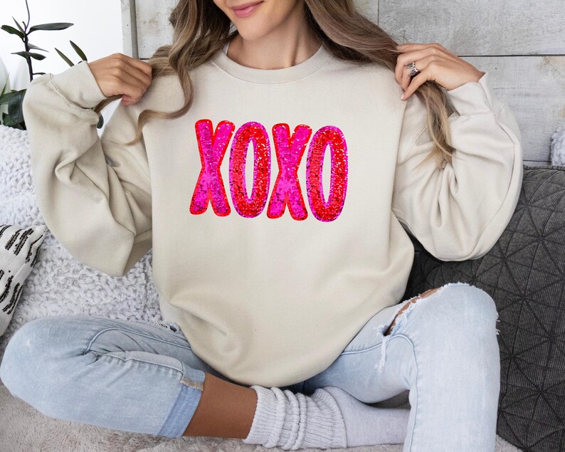 以下が含まれることがあります： クリーム色のスウェットシャツに、ピンクと赤のグリッターで「XOXO」のグラフィックが施されています。