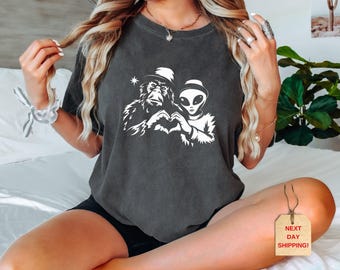 May include: A dark gray t-shirt with a white graphic of a monkey and an alien holding hands. The monkey is wearing a hat and the alien has a large head.