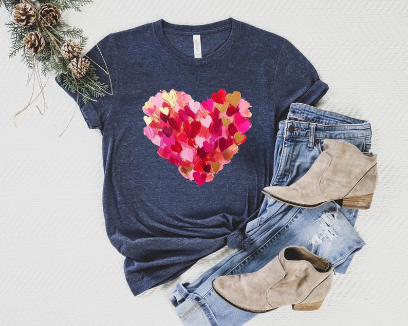 Op de afbeelding: Een marineblauw t-shirt met een groot roze en gouden hartontwerp dat is gemaakt van kleinere harten. Het hart is gecentreerd op het shirt.