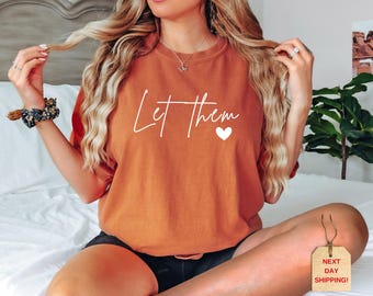 May include: A burnt orange t-shirt with the words "Let them" in white script and a white heart. The shirt is a short-sleeved style. The model is wearing black shorts and a floral scrunchie.