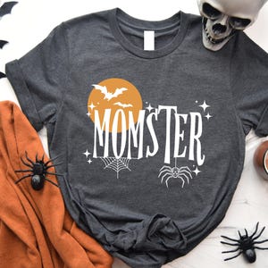 May include: A dark gray t-shirt with a white graphic design. The design features a yellow moon with bats flying around it, a spider web, a spider, and the text "MOMSTER".