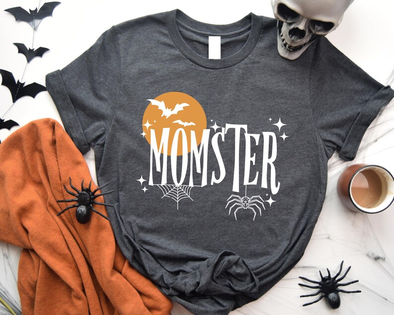 May include: A dark gray t-shirt with a white graphic design. The design features a yellow moon with bats flying around it, a spider web, a spider, and the text "MOMSTER".