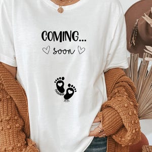 Op de afbeelding: Een wit t-shirt met zwarte tekst die COMING... soon leest met twee kleine hartjes. Onder de tekst staat een afbeelding van twee babyvoetjes met hartjes in de hielen en de tekst DECEMBER 2023 gedrukt tussen de voetjes in.