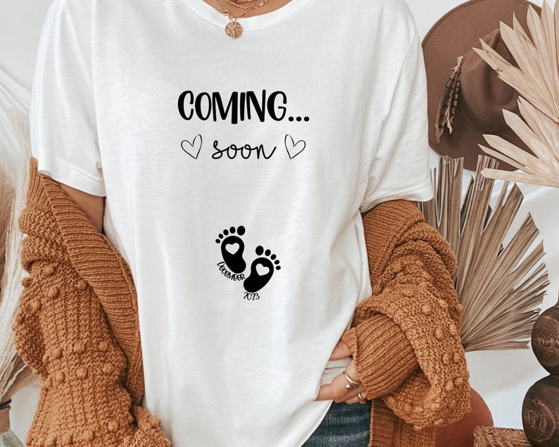 Op de afbeelding: Een wit t-shirt met zwarte tekst die COMING... soon leest met twee kleine hartjes. Onder de tekst staat een afbeelding van twee babyvoetjes met hartjes in de hielen en de tekst DECEMBER 2023 gedrukt tussen de voetjes in.