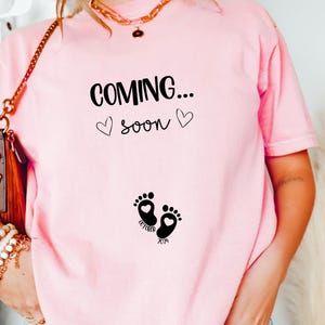 Op de afbeelding: Een lichtroze t-shirt met de zwarte tekst "COMING... soon" en twee kleine hartjes. Onder de tekst staan twee zwarte babyvoetjes, elk met een klein hartje erin, en de woorden "OCTOBER" en "2024" zijn bij de voetjes gedrukt.