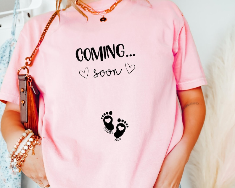 Op de afbeelding: Een lichtroze t-shirt met de zwarte tekst "COMING... soon" en twee kleine hartjes. Onder de tekst staan twee zwarte babyvoetjes, elk met een klein hartje erin, en de woorden "OCTOBER" en "2024" zijn bij de voetjes gedrukt.