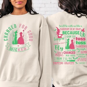 May include: A cream-colored sweatshirt with a green and pink graphic of two witches and the text "Changed for Good Wicked". The back of the sweatshirt has green text that reads "You'll be with me like a handprint on my heart, because I knew you everyone deserves the chance to fly toss toss dancing through life I have been shiz changed for good I think I'll try defying gravity you're gonna be popular as long as you're mine".
