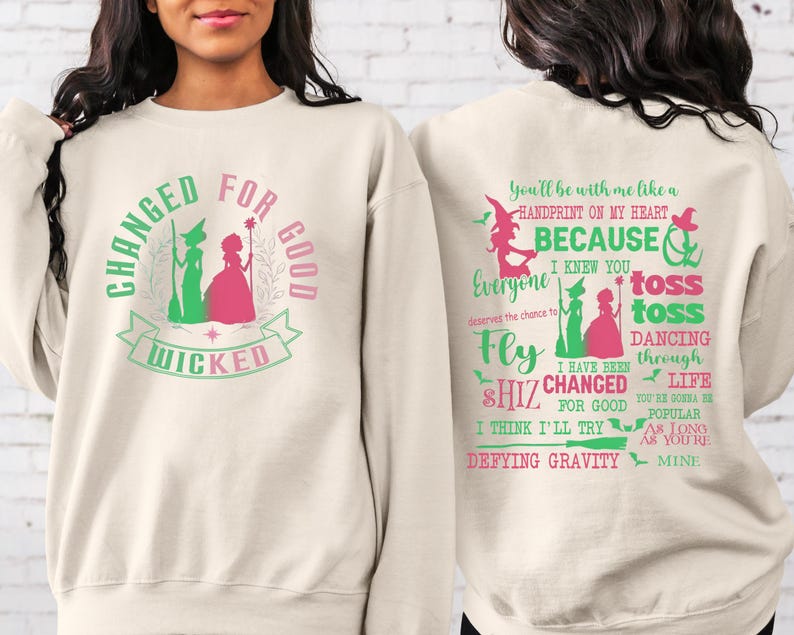 May include: A cream-colored sweatshirt with a green and pink graphic of two witches and the text "Changed for Good Wicked". The back of the sweatshirt has green text that reads "You'll be with me like a handprint on my heart, because I knew you everyone deserves the chance to fly toss toss dancing through life I have been shiz changed for good I think I'll try defying gravity you're gonna be popular as long as you're mine".
