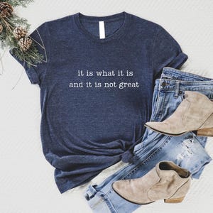 May include: Navy blue t-shirt with the text "it is what it is and it is not great" in white. The shirt is paired with blue jeans and tan ankle boots. A decorative pine branch with pinecones is in the upper left corner.