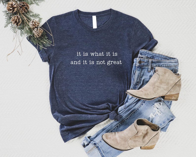 May include: Navy blue t-shirt with the text "it is what it is and it is not great" in white. The shirt is paired with blue jeans and tan ankle boots. A decorative pine branch with pinecones is in the upper left corner.