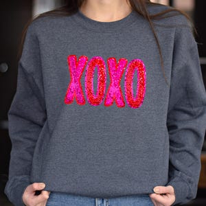 以下が含まれることがあります： ピンクと赤のグリッターで XOXO とデザインされたダークグレーのスウェットシャツです。