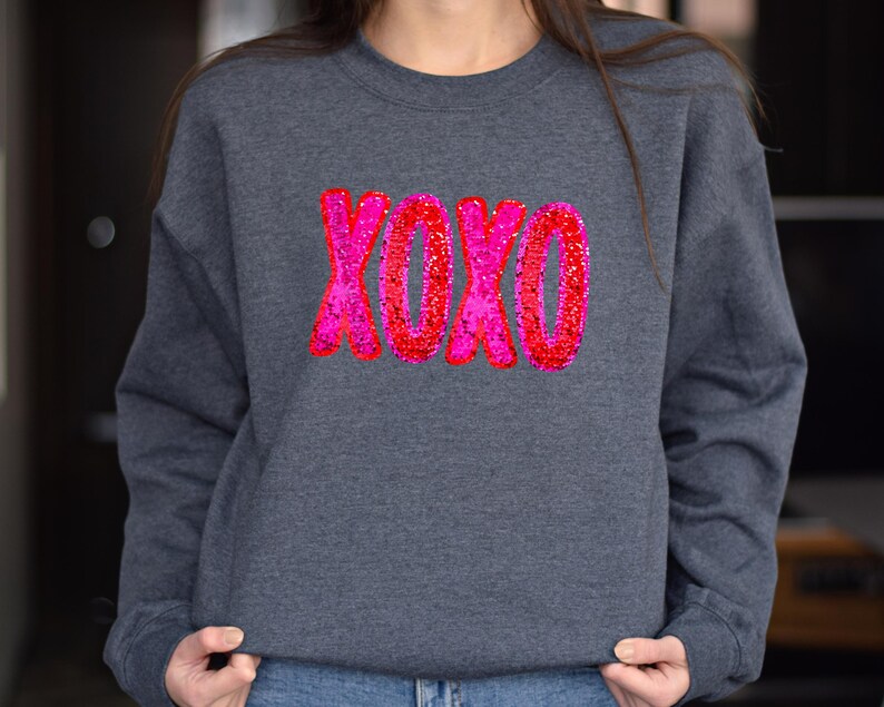 以下が含まれることがあります： ピンクと赤のグリッターで XOXO とデザインされたダークグレーのスウェットシャツです。