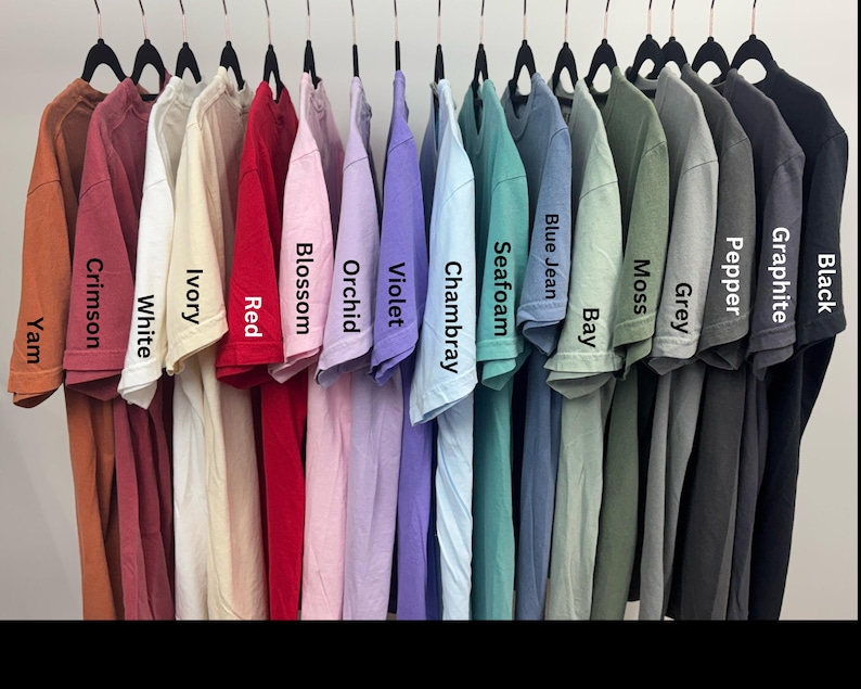 Puede incluir: Una colecci&oacute;n de camisetas de varios colores colgadas de perchas negras. Los colores incluyen Yam, Crimson, White, Ivory, Red, Blossom, Orchid, Violet, Chambray, Seafoam, Blue Jean, Bay, Moss, Grey, Pepper, Graphite y Black.