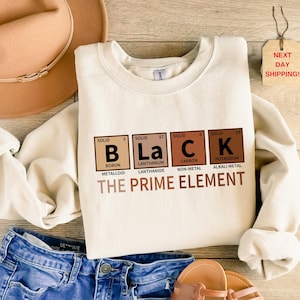May include: Cream-colored sweatshirt with the word "BLACK" spelled out using periodic table elements. The text "THE PRIME ELEMENT" is below the word. The sweatshirt is paired with blue jeans and a brown hat.