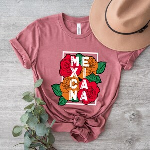 May include: A dusty rose t-shirt featuring the word "MEXICANA" in a white frame, with colorful roses in red, orange, and green. A tan hat rests on the shirt. The shirt is knotted at the bottom.