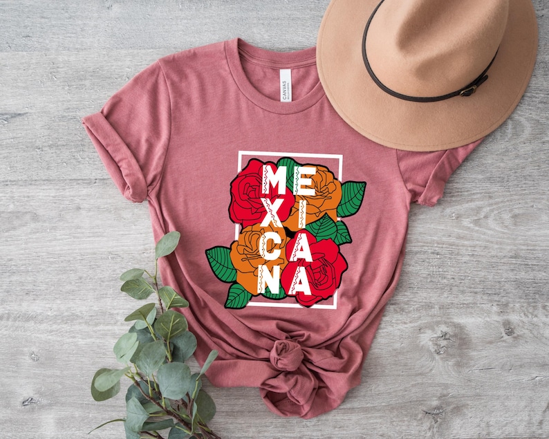 May include: A dusty rose t-shirt featuring the word "MEXICANA" in a white frame, with colorful roses in red, orange, and green. A tan hat rests on the shirt. The shirt is knotted at the bottom.