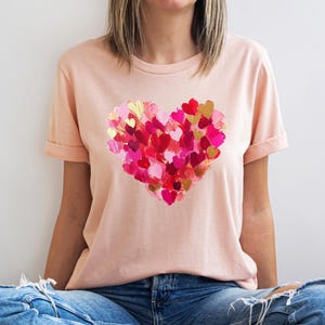 Op de afbeelding: Een lichtroze T-shirt met een groot hartontwerp gemaakt van veel kleinere harten in tinten roze, rood en goud.