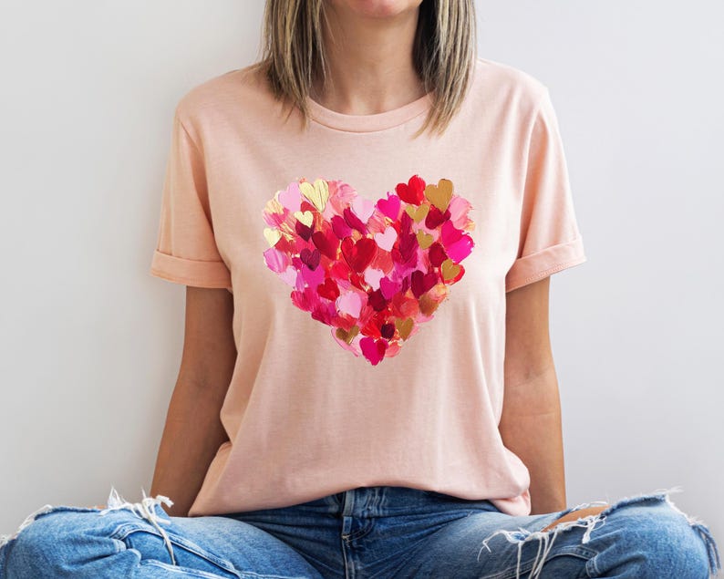 Op de afbeelding: Een lichtroze T-shirt met een groot hartontwerp gemaakt van veel kleinere harten in tinten roze, rood en goud.