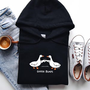May include: Black hooded sweatshirt with a white goose design and the text "Goose Bumps". The sweatshirt is laid flat on a white surface with a pair of gray sneakers and blue jeans.