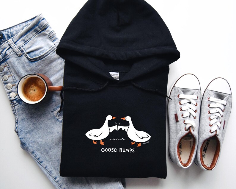 May include: Black hooded sweatshirt with a white goose design and the text "Goose Bumps". The sweatshirt is laid flat on a white surface with a pair of gray sneakers and blue jeans.