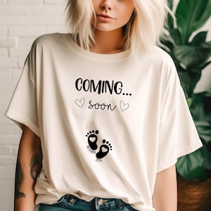 Op de afbeelding: Een cr&egrave;mekleurig t-shirt met korte mouwen en zwarte tekst COMING... soon met twee kleine hartjes. Onder de tekst staan twee zwarte babyvoetjes met hartvormige centra en de woorden October 2024 eronder gedrukt.