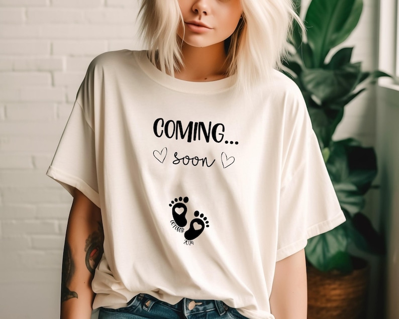Op de afbeelding: Een cr&egrave;mekleurig t-shirt met korte mouwen en zwarte tekst COMING... soon met twee kleine hartjes. Onder de tekst staan twee zwarte babyvoetjes met hartvormige centra en de woorden October 2024 eronder gedrukt.