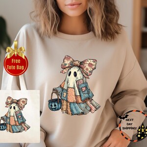 May include: Beige sweatshirt featuring a whimsical ghost design with a patchwork quilt-style outfit and a large bow. The ghost holds a blue jack-o'-lantern bucket. A tote bag with the same ghost design is also visible. The words "Free Tote Bag" are on a gold-ribboned tag.