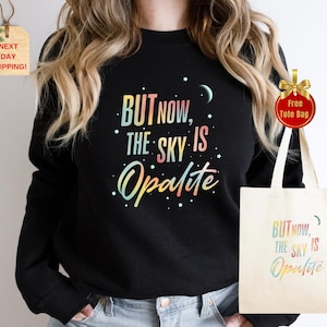 May include: Black sweatshirt with the text "BUT NOW, THE SKY IS Opalite" in a colorful, rainbow-like font. A matching tote bag is visible. The design includes stars and a crescent moon.