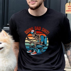 May include: Black t-shirt with a graphic of a skeleton on a toilet, wearing a baseball cap and sneakers. The text "DOING DAD SHIT!" is arched above the image in red and orange. A small dog is in the image.