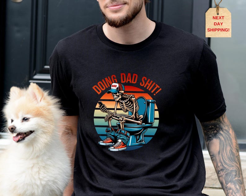 May include: Black t-shirt with a graphic of a skeleton on a toilet, wearing a baseball cap and sneakers. The text "DOING DAD SHIT!" is arched above the image in red and orange. A small dog is in the image.