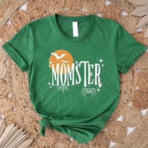 May include: Green t-shirt with a white graphic of a spider, a crescent moon, and bats. The text "MOMSTER" is written in a white, stylized font.