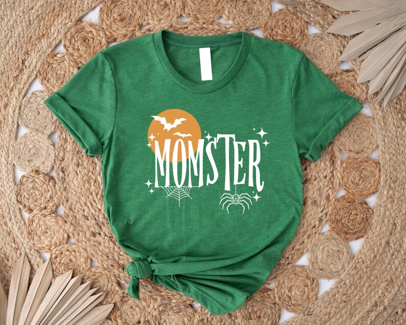 May include: Green t-shirt with a white graphic of a spider, a crescent moon, and bats. The text "MOMSTER" is written in a white, stylized font.