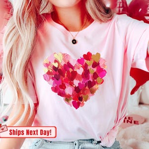 Op de afbeelding: Roze T-shirt met een groot hartontwerp gemaakt van veel kleine, kleurrijke hartjes. Het hart is geschilderd in tinten roze, rood en goud.