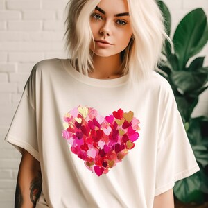 Op de afbeelding: Een wit T-shirt met een groot roze, rood en gouden hartontwerp gemaakt van kleinere hartjes. Het hartontwerp is gecentreerd op het shirt.