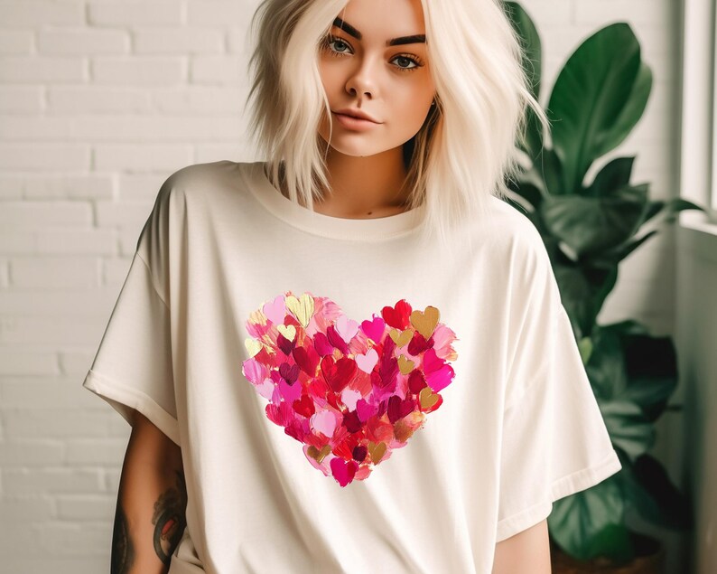 Op de afbeelding: Een wit T-shirt met een groot roze, rood en gouden hartontwerp gemaakt van kleinere hartjes. Het hartontwerp is gecentreerd op het shirt.