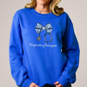 May include: Royal blue sweatshirt with a white bow and stethoscope design. The text "Respiratory Therapist" is printed below the design.