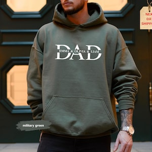 May include: Military green hoodie with the word "DAD" in white block letters, with "EMMA & OLIVIA & LIAM" underneath. The hoodie has a front pocket and a hood. The image also includes a tag that says "NEXT DAY SHIPPING!"