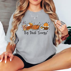 Puede incluir: Una camiseta gris claro con un gr&aacute;fico de alimentos de oto&ntilde;o, que incluye un caf&eacute; con leche de calabaza, tarta, pavo y magdalena. El texto "Big Back Energy" est&aacute; debajo de las ilustraciones de comida. La camiseta es de estilo casual.