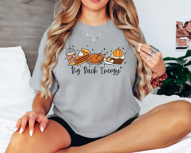 Puede incluir: Una camiseta gris claro con un gr&aacute;fico de alimentos de oto&ntilde;o, que incluye un caf&eacute; con leche de calabaza, tarta, pavo y magdalena. El texto "Big Back Energy" est&aacute; debajo de las ilustraciones de comida. La camiseta es de estilo casual.
