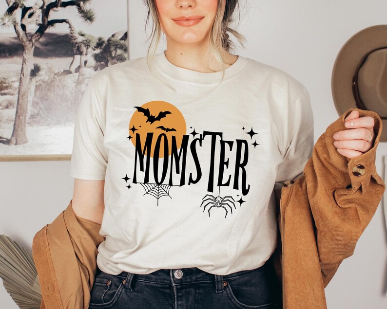 May include: A white t-shirt with a black graphic design that says "Momster" with a spider, bats, and a moon.